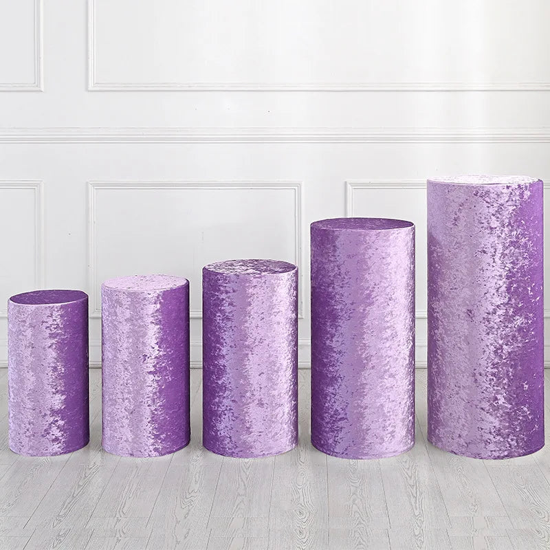 A row of four cylindrical spandex stands in varying heights and a purple glitter fabric, displayed against a white paneled wa