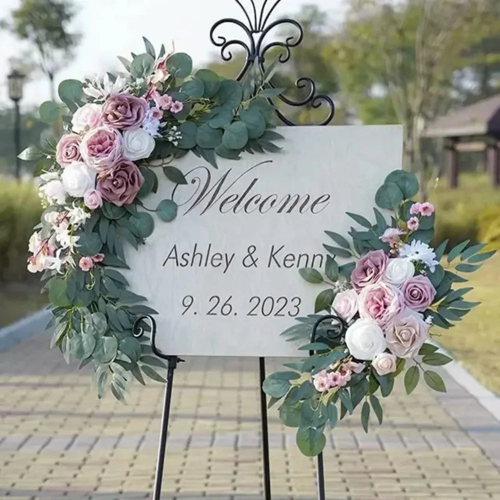A white welcome sign decorated with dusty rose and white flowers and green eucalyptus leaves for a wedding.