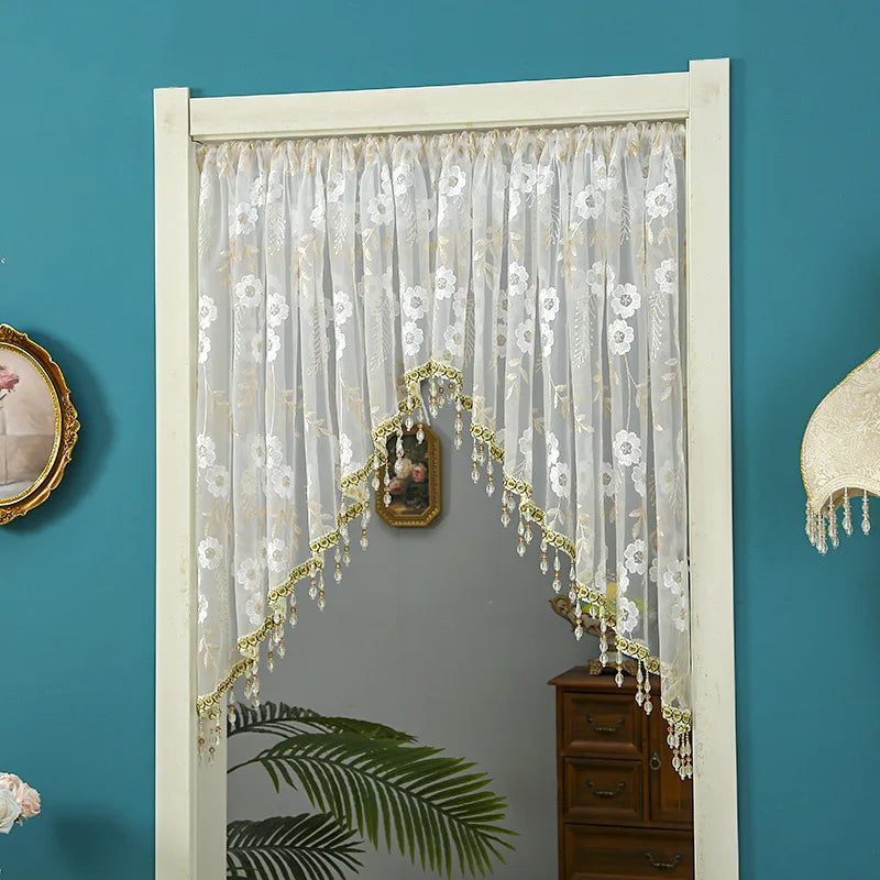 A sheer lace door curtain with gold tassels hangs in a white frame against a teal wall.