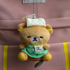 A fluffy brown Rilakkuma bear plush toy with a green collar is hanging from a keychain on a pink fabric bag.