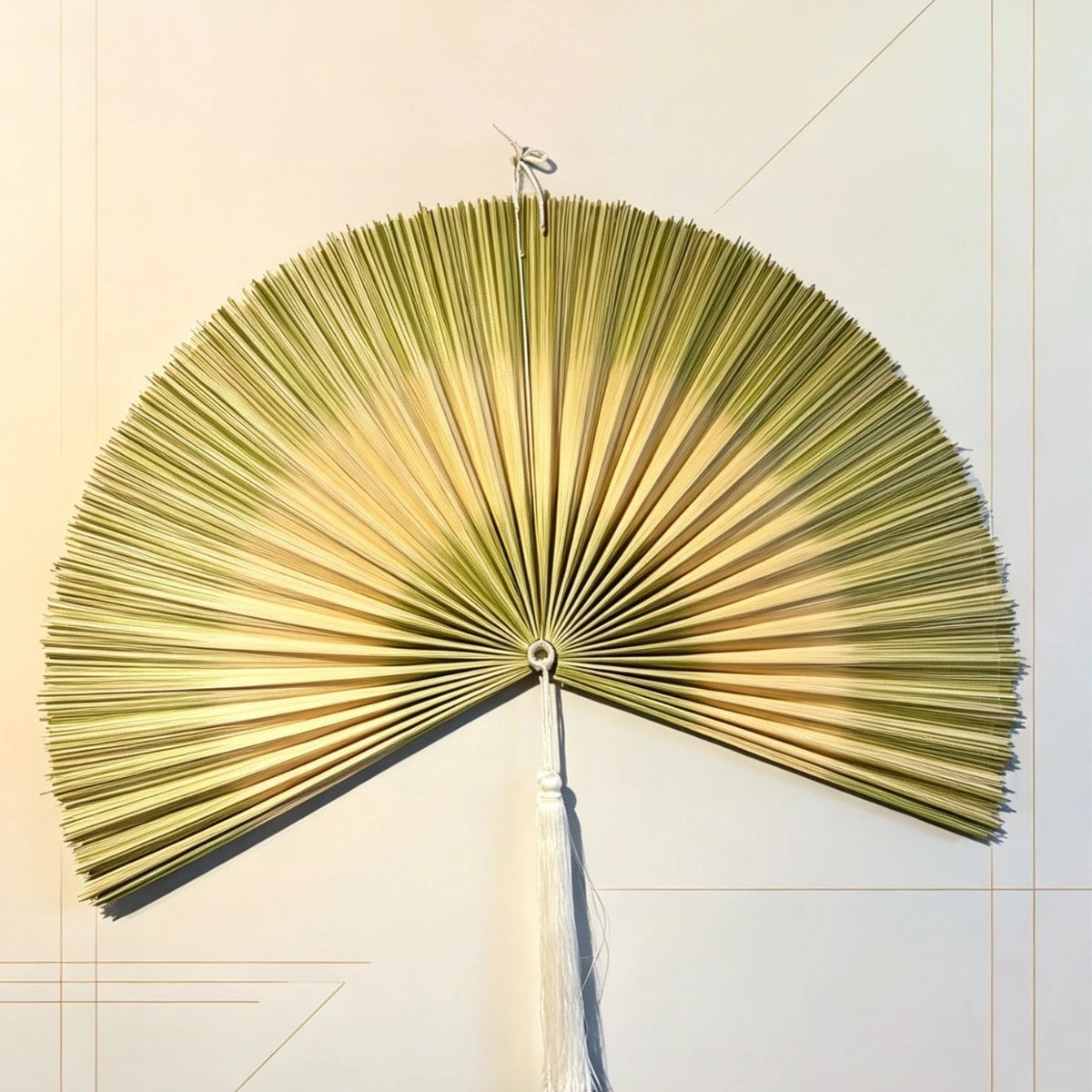 A handwoven bamboo wall fan decor with a golden-green color and a white tassel, displayed against a light background.