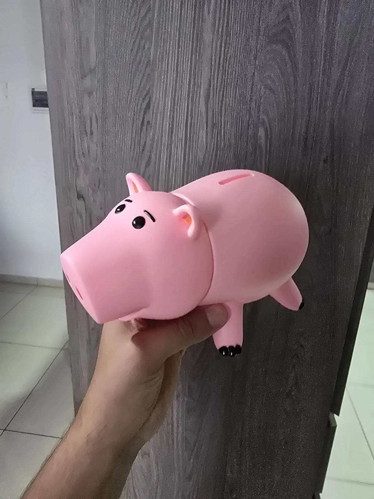 A hand holds a pink piggy bank with a cartoon face against a gray wooden surface.