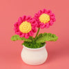 A hand-woven crochet flower in vibrant pink and yellow sits in a white pot.