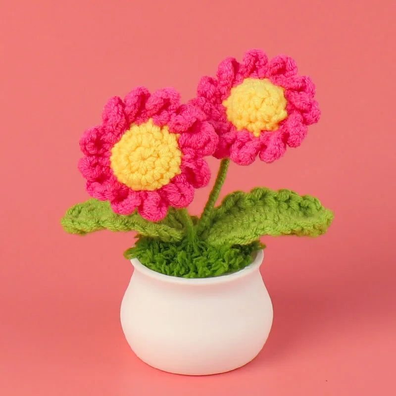 A hand-woven crochet flower in vibrant pink and yellow sits in a white pot.