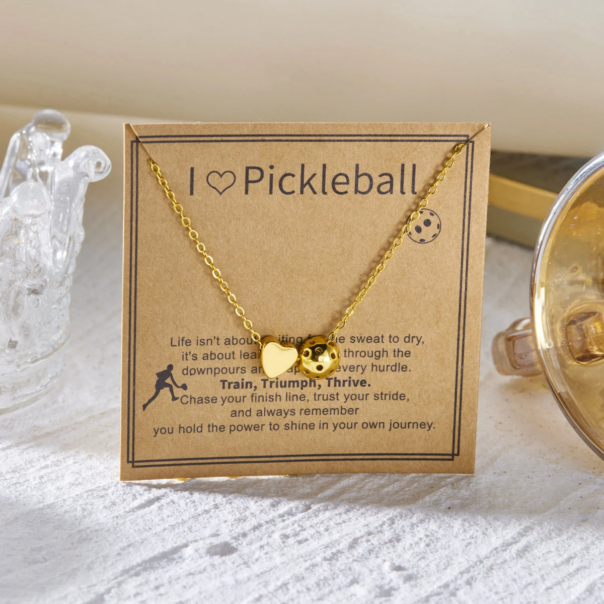 Gold necklace with a pickleball charm and inspirational text on a kraft paper card
