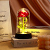 A glass dome containing a red rose with glowing yellow leaves, displayed on a dark base.