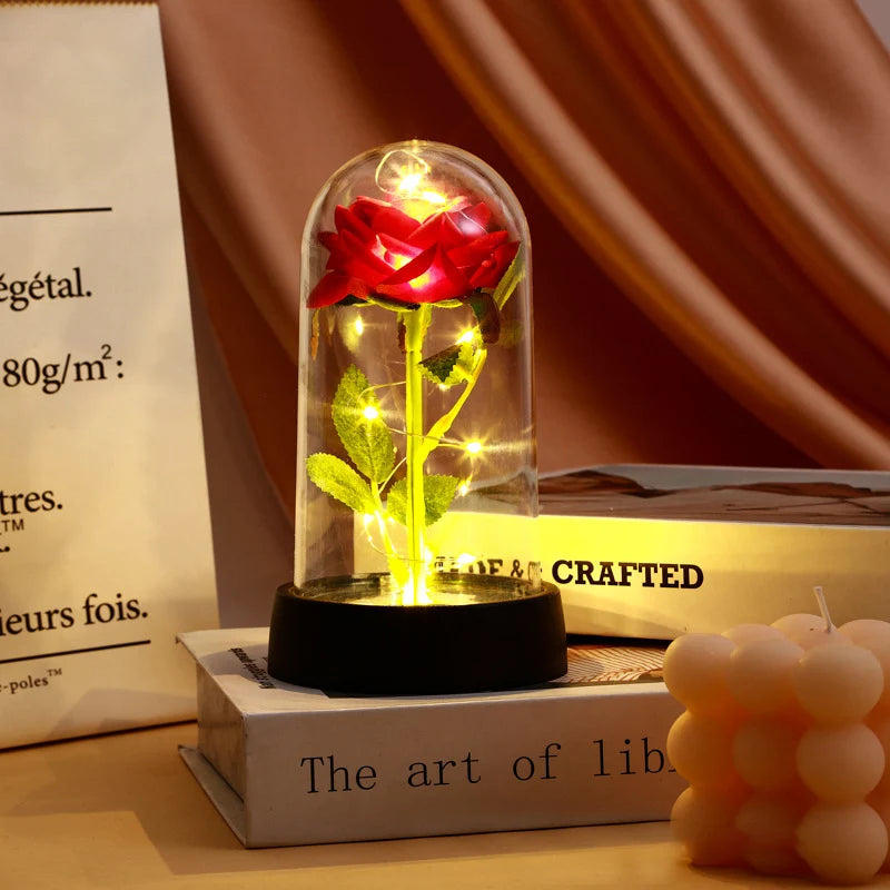 A glass dome containing a red rose with glowing yellow leaves, displayed on a dark base.