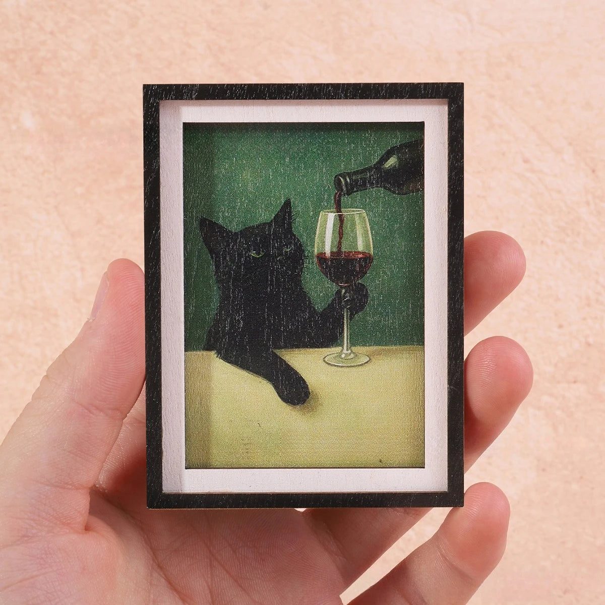 Gerahmtes Foto einer schwarzen Katze, die Rotwein aus einem Glas trinkt, gehalten von einer Hand.