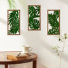 Three framed pieces of green tropical leaf wall art on a white wall next to a wooden table.