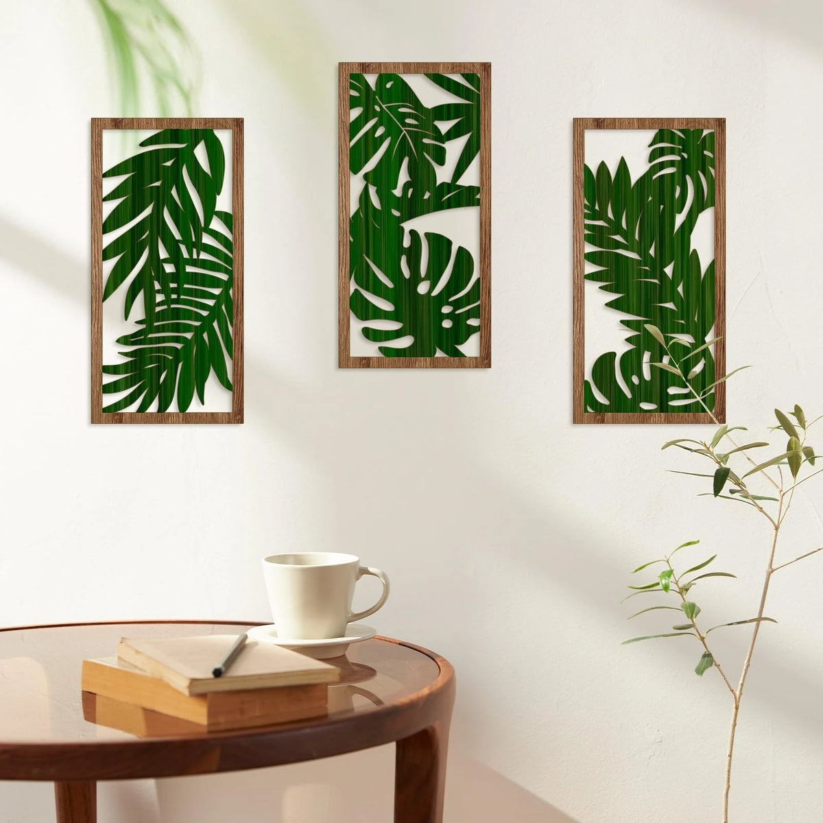 Three framed pieces of green tropical leaf wall art on a white wall next to a wooden table.