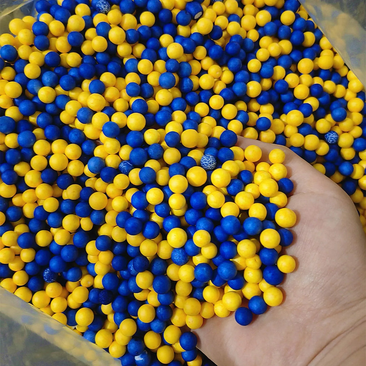 A hand is holding a large pile of yellow and blue pastel foam beads for crafting.