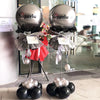 Two silver balloons displayed on an adjustable metal column stand with black and red ribbon accents.