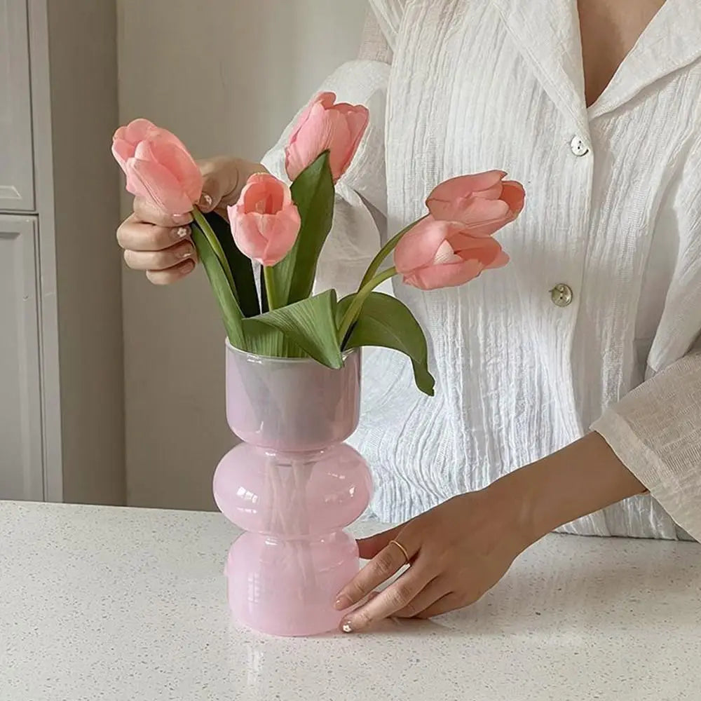 A hand holds a pink glass flower vase filled with light pink tulips on a white countertop.