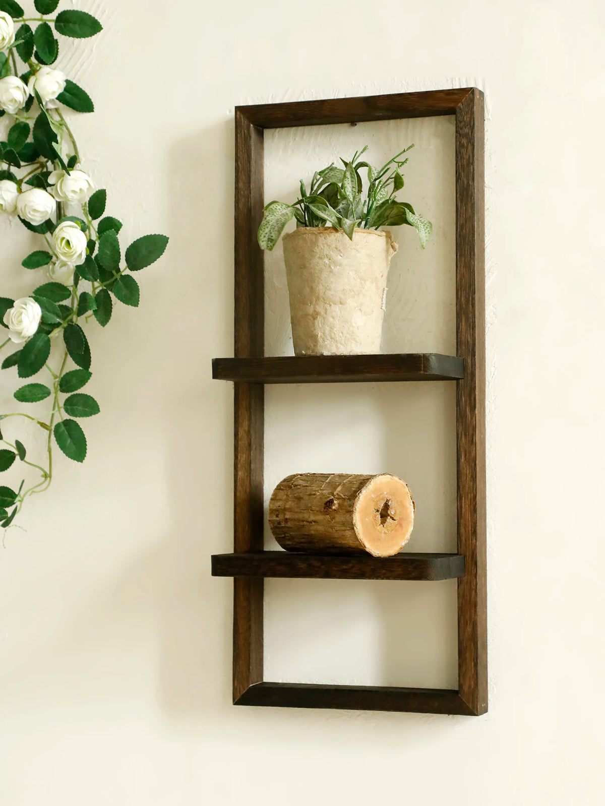 A dark wood floating wall shelf with two tiers displaying a potted plant and a wooden log.