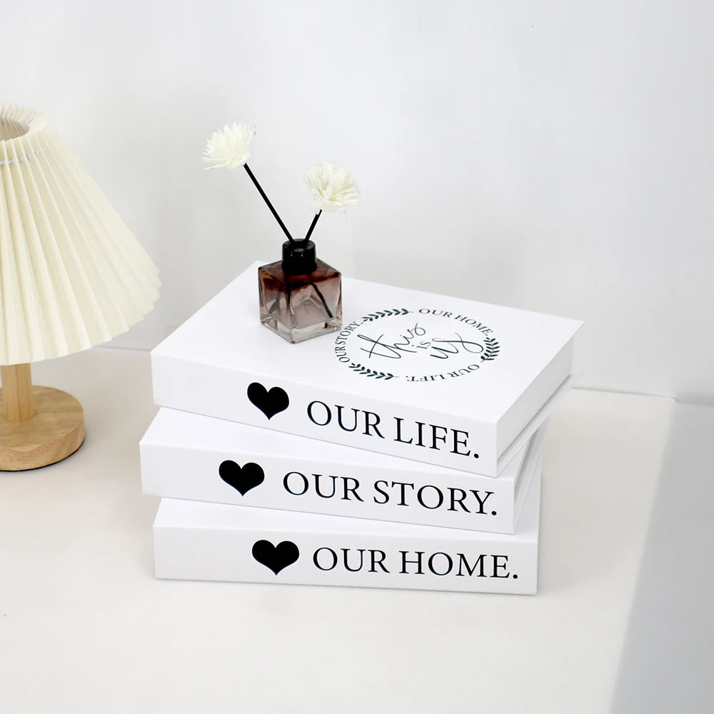 A set of three white decorative fake books with black hearts and text on them, displayed next to a white lamp.
