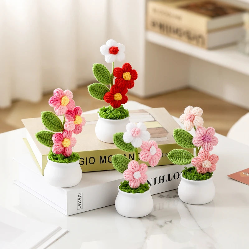 Hand woven crochet flowers in pink and white are potted in green moss and displayed on a white surface.