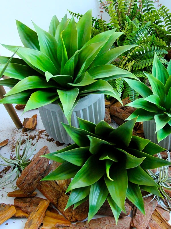 A vibrant green artificial agave plant in a gray pot surrounded by natural wood pieces and ferns.