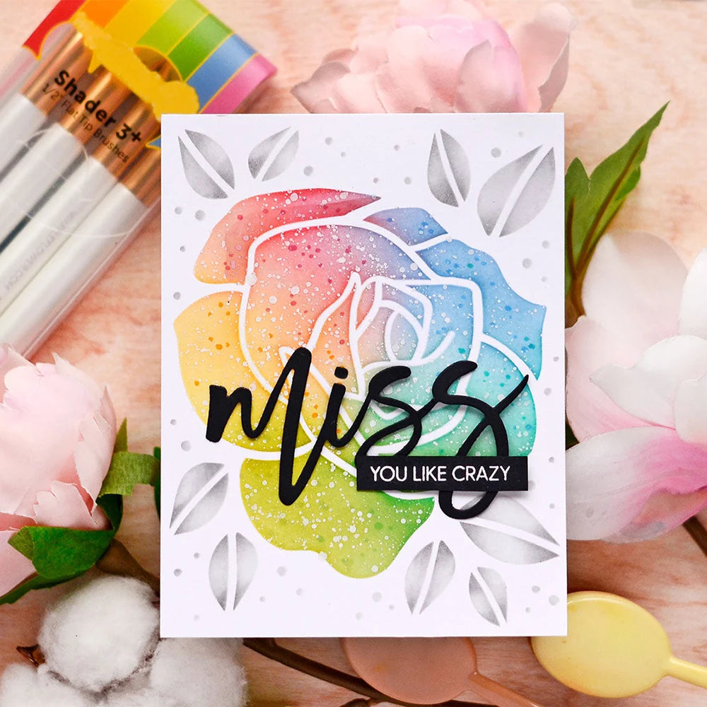 A card featuring the words Miss You Like Crazy surrounded by floral stencils and stamps on a pink background.