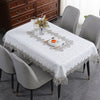 A white tablecloth with embroidered lace trim is set on a dining table surrounded by modern gray chairs.