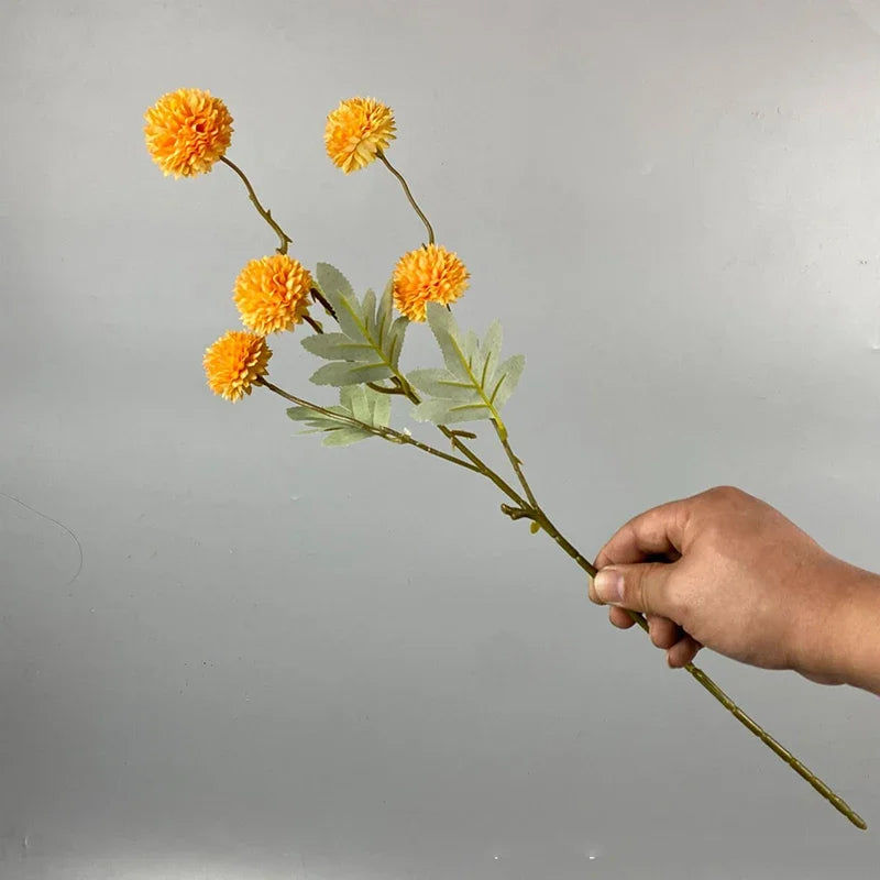 Una mano sostiene un tallo con varias flores artificiales de margarita de chrysanthemum amarillo brillante y hojas verdes con