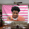 A young man with dreadlocks wearing an orange shirt is centered against a pink and white striped flag banner.
