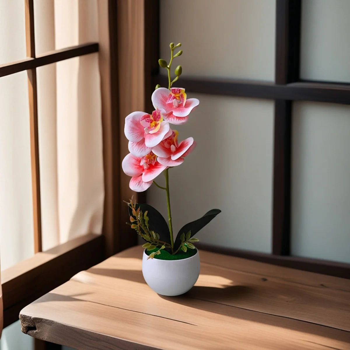 A realistic artificial pink orchid plant in a white pot sits on a wooden table next to a window.