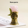 A rose quartz crystal is displayed in a brass bell, featuring a natural copper finish and green leaves.