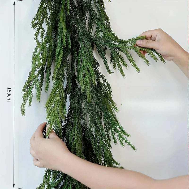 A hand is holding a long, green artificial Norfolk pine garland with warm white lights against a white background.