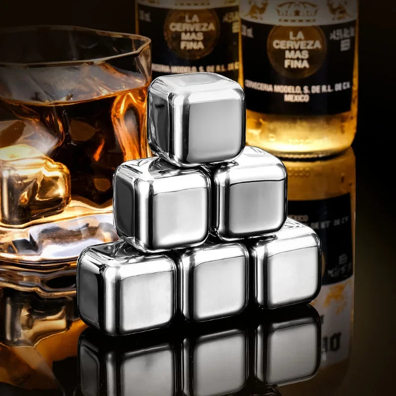 A stack of reusable stainless steel ice cubes resting on a reflective surface next to a glass of amber liquid and bottles of 