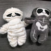 A white mummy skeleton plush toy next to a gray mummy skeleton plush toy.