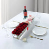 A clear acrylic rectangular vase filled with red roses and pink flowers on a white table with wine and dinnerware.