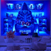 A person sits in a gaming room surrounded by UV reactive light and displays featuring game imagery, holding a controller.