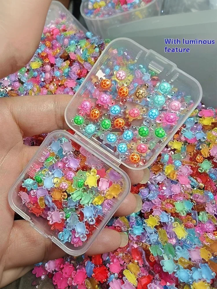A hand holds an open clear plastic container filled with colorful glow-in-the-dark resin turtle figurines for crafts.
