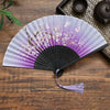 A vintage silk folding fan with purple and white floral patterns is displayed on a wooden surface.