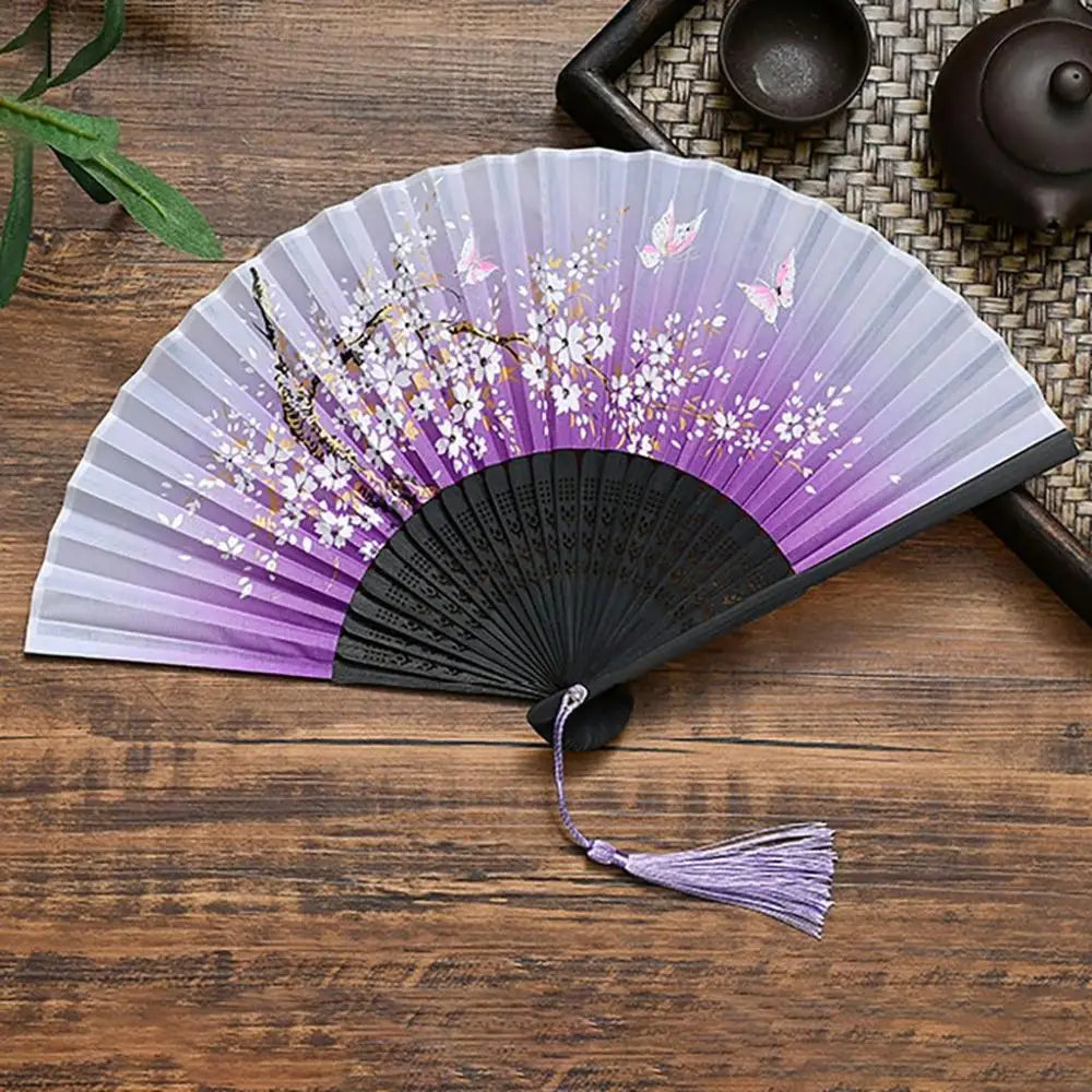 A vintage silk folding fan with purple and white floral patterns is displayed on a wooden surface.