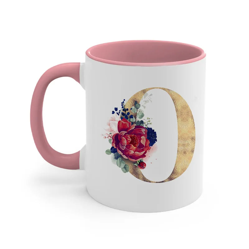 A pink ceramic coffee mug featuring a large gold letter O decorated with colorful floral accents.