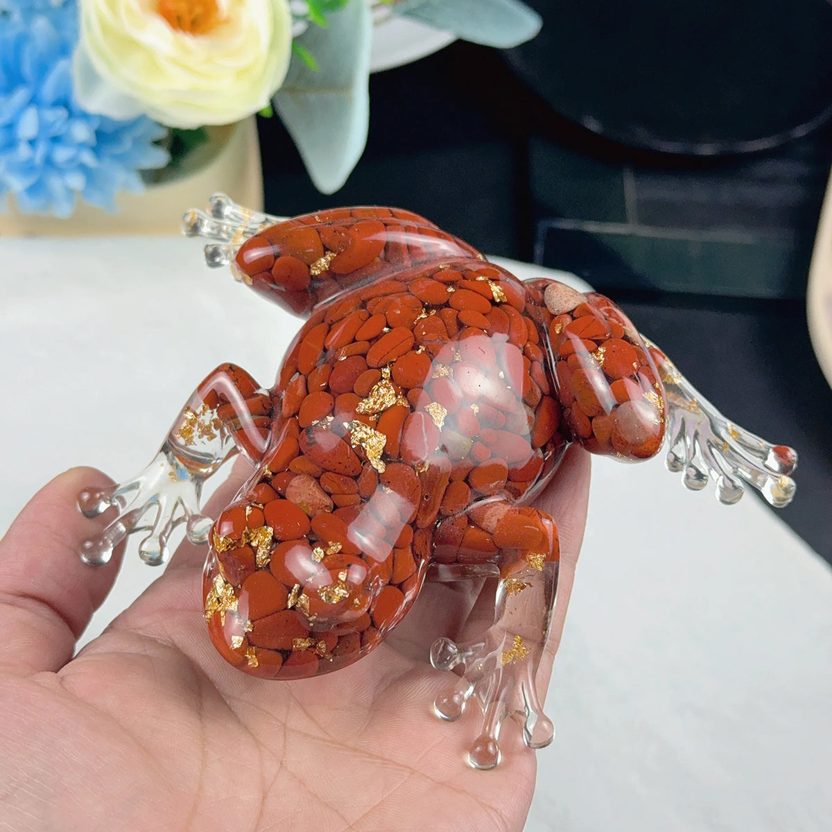 A hand holds a resin craft decoration shaped like an orange-brown frog with clear limbs and gold accents.