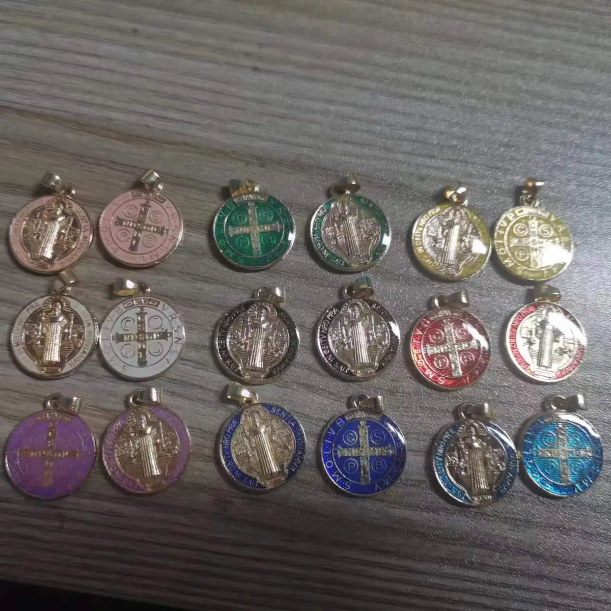A collection of twelve double-sided St. Benedict medals in various colors and finishes arranged on a surface.