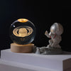 A glass crystal ball night light featuring a solar system design on a wooden base next to a small white figurine.