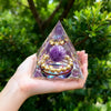 Handmade resin orgonite pyramid with purple natural obsidian and a copper coil set against a green outdoor background