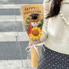 A handcrafted crochet flower bouquet featuring a yellow sunflower and a small teddy bear wearing a graduation cap.