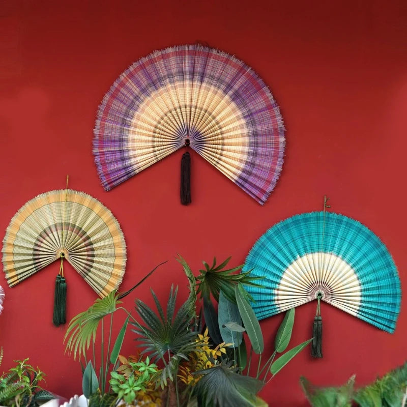 A handwoven bamboo wall fan decor with a rainbow gradient design mounted on a red wall.