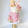 A tiered cake decorated with pink and gold elements, featuring a bear topper and floral accents.