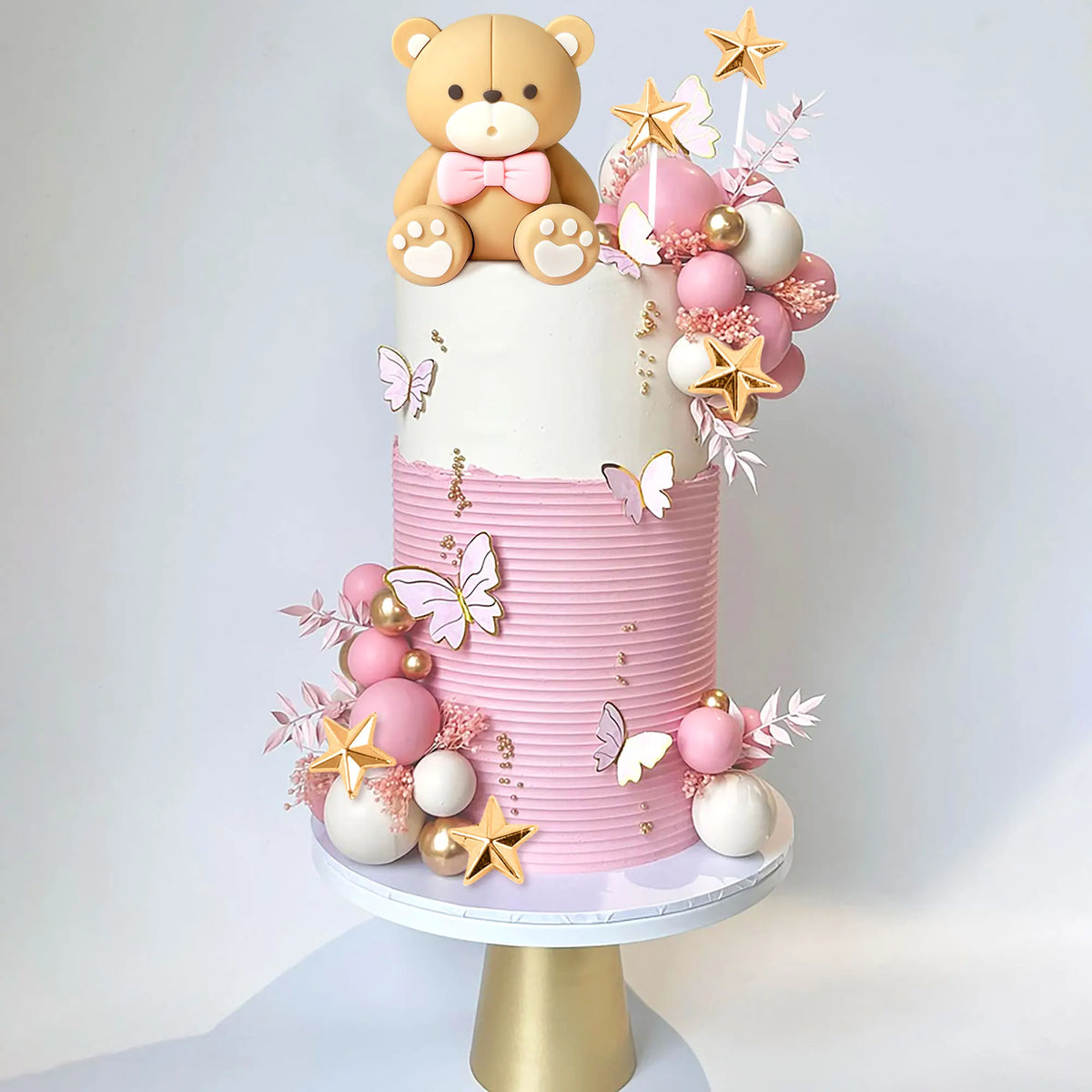 A tiered cake decorated with pink and gold elements, featuring a bear topper and floral accents.