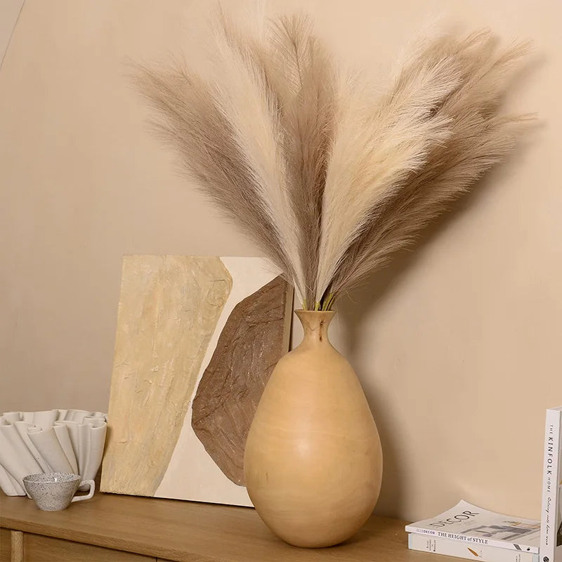 A vase holds dried artificial pampas grass branches against a light beige wall with wooden decor.