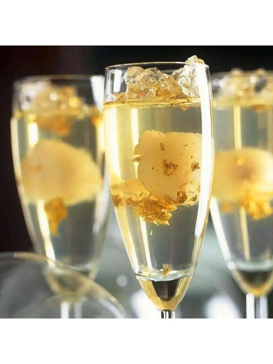 A close-up of a champagne flute filled with sparkling liquid and edible gold leaf flakes for decoration.