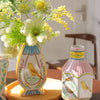 A pink and white ceramic bird floral vase with blue accents sits on a wooden table next to green foliage.
