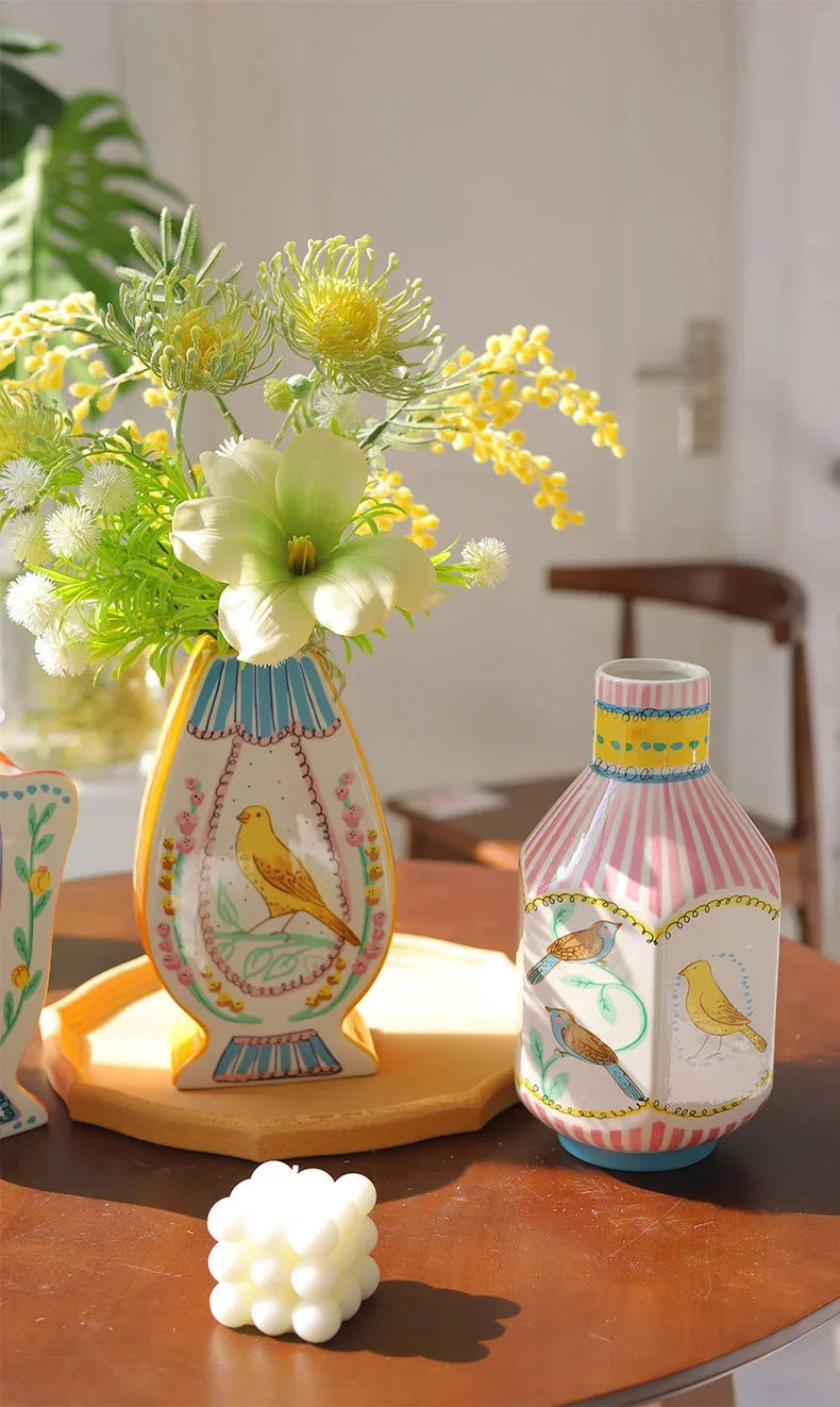A pink and white ceramic bird floral vase with blue accents sits on a wooden table next to green foliage.