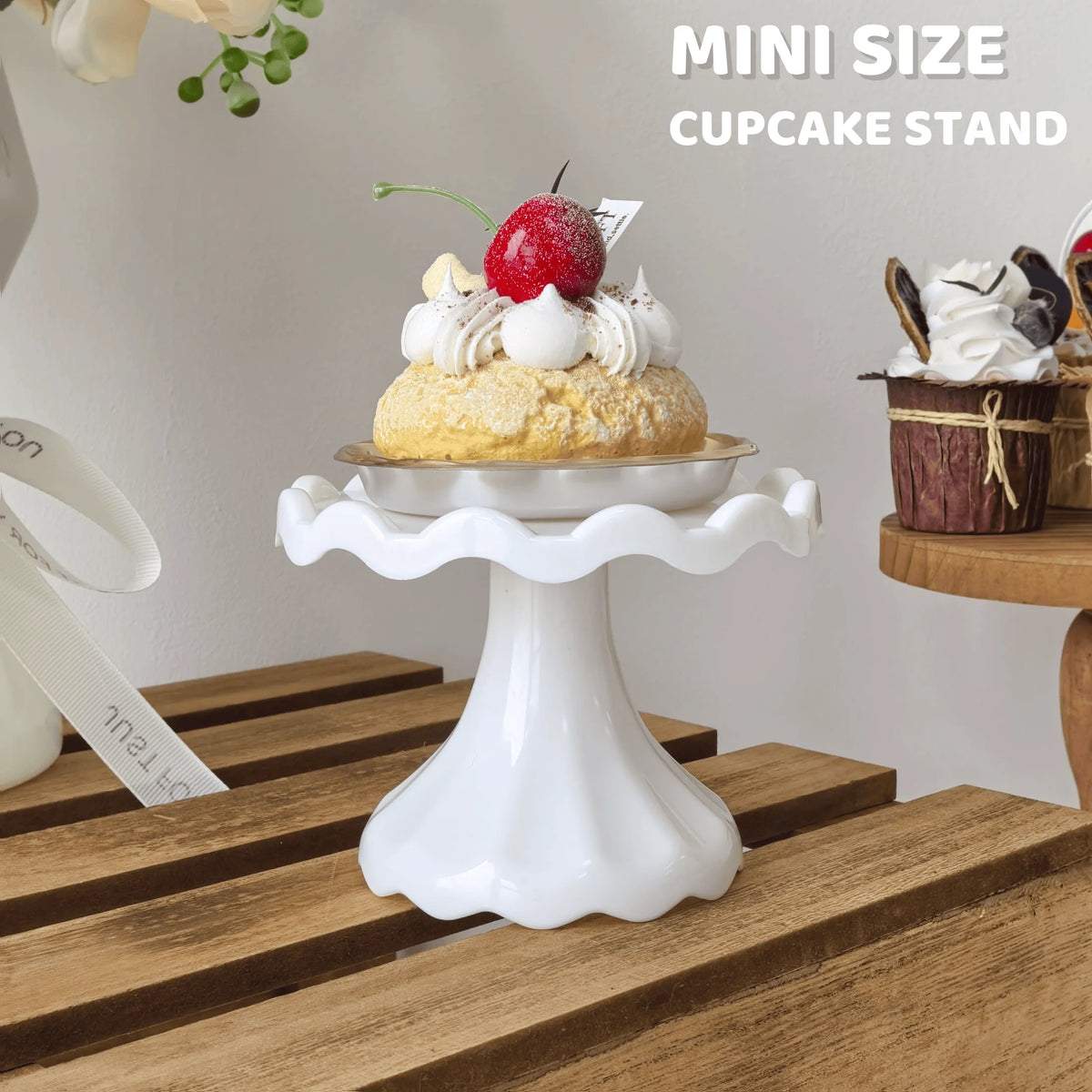 A white mini cupcake stand displaying a single cupcake topped with whipped cream and a red berry on a wooden surface.