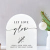 A white acrylic sign with black script that reads Let Love Glow and instructs to grab a glow stick for a dance floor.